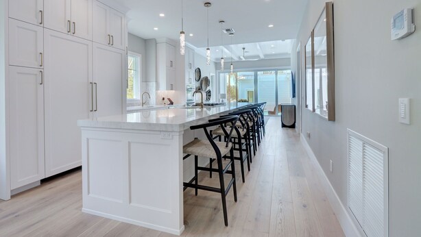 Una cocina moderna con armarios blancos, una isla central con asientos tipo barra, iluminación colgante y vistas al exterior.