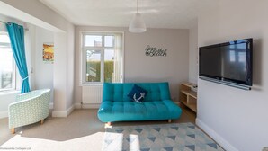 Ein gemütliches Wohnzimmer mit einem türkisfarbenen Sofa, einem kleinen Beistelltisch, einem an der Wand montierten Fernseher und einem Fenster mit Vorhängen.