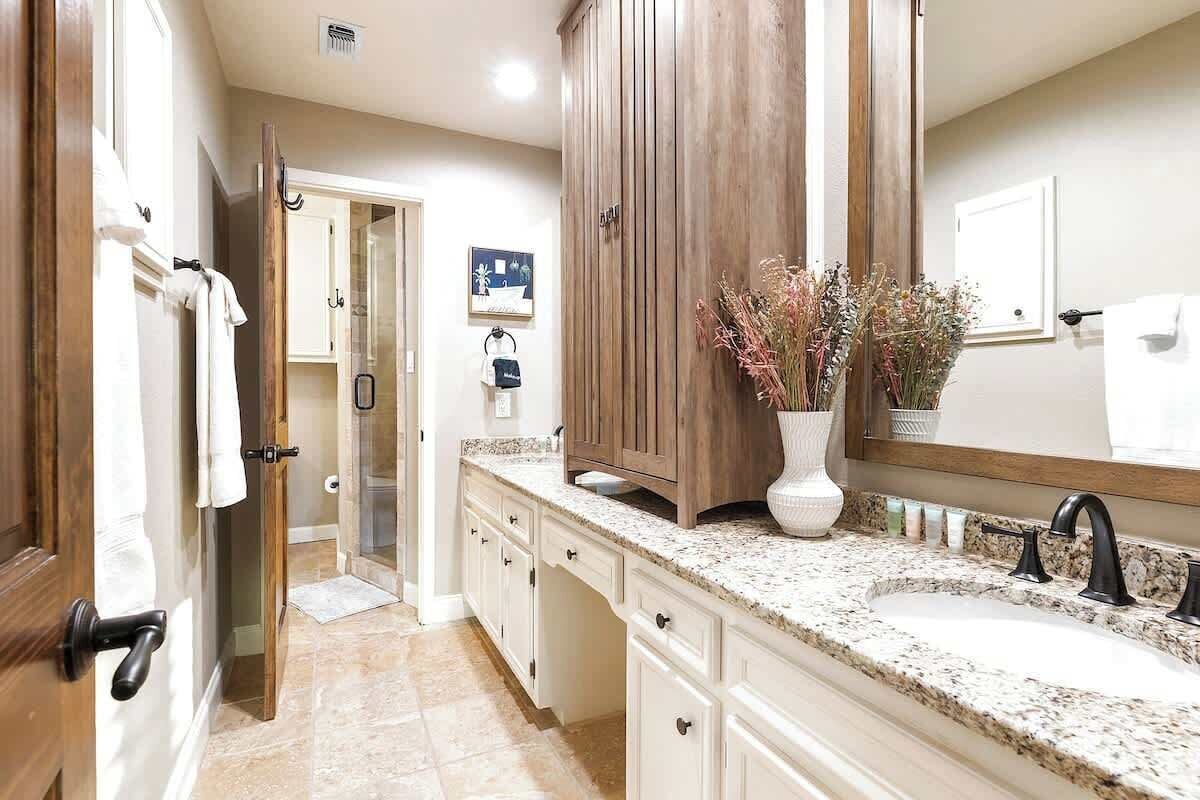 A bathroom with a granite countertop, a large mirror, wooden cabinets, and a vase with dried flowers.