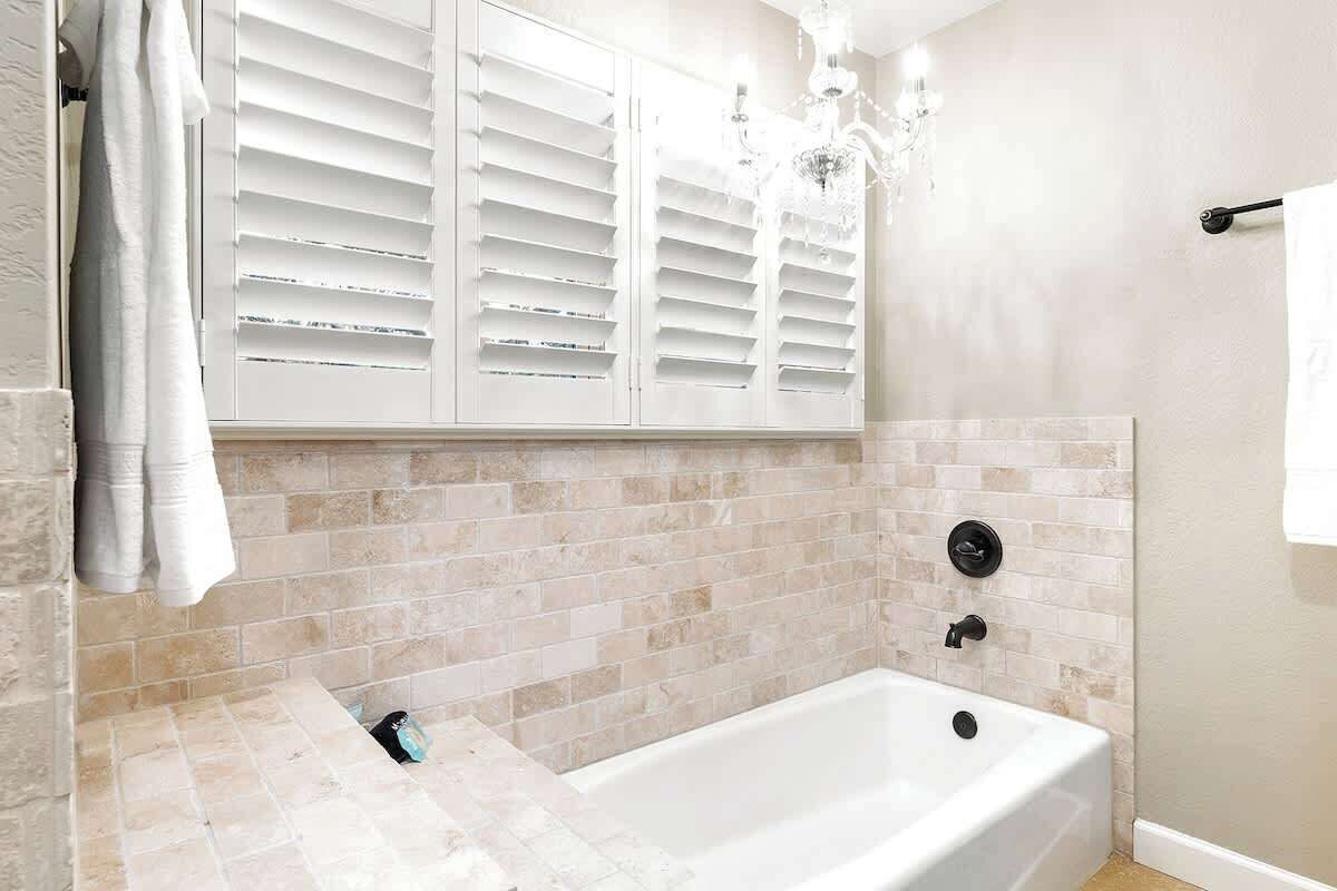 A bathroom with a bathtub, white tiled walls, a white window with shutters, and a chandelier.