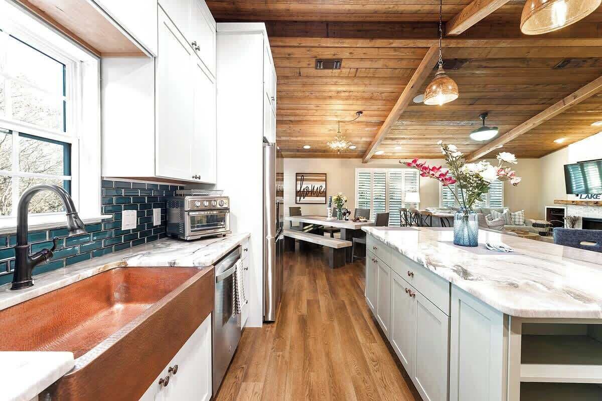 Una cucina con mobili bianchi, un lavello in rame, uno schienale in mattoni e travi al soffitto in legno.