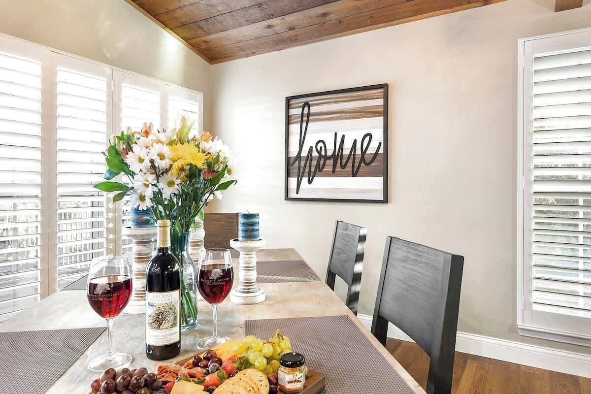 Una mesa de comedor con una botella de vino, copas, una bandeja de frutas y un jarrón de flores.