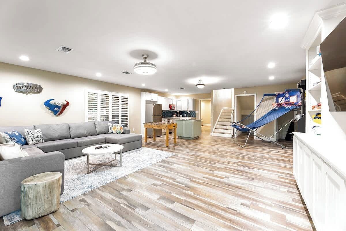 A spacious living room with a grey sofa, a coffee table, and a wooden floor. There is a kitchen area with a dining table and a staircase leading to an upper level.