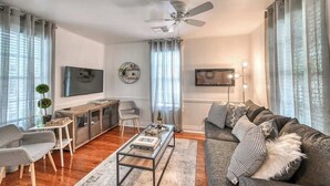 A modern living room with a sofa, coffee table, TV, and a ceiling fan. - Charming Cottage Near Main With Patio&firepit!! (Fredericksburg)