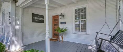 A porch with a swing chair, a potted plant, and a sign reading "The Pottery Barn Cottage."