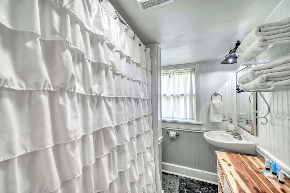 Ein Badezimmer mit weißer, geraffter Duschvorhang, einem Holzkosmetikschrank mit Waschbecken und einem Spiegel.