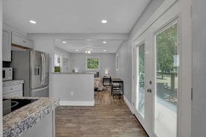 A modern kitchen with a central island, a dining area with a table, and a large window with blinds. - Grey Haus Casita-peaceful Hill Country Views! (Fredericksburg)