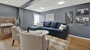 A modern living room with a sectional sofa, a coffee table, and a seating area with chairs. There is a built-in grill and a large window providing natural light.