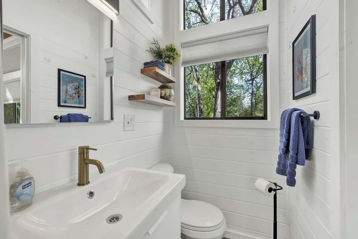 Une salle de bain avec un lavabo blanc, des toilettes et une fenêtre donnant sur des arbres. On y trouve des étagères avec des plantes et un tableau encadré accroché au mur.