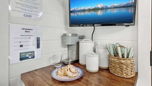 A hotel room with a flat-screen TV displaying a mountain landscape, a coffee maker, a glass of water, and a plate with pastries.