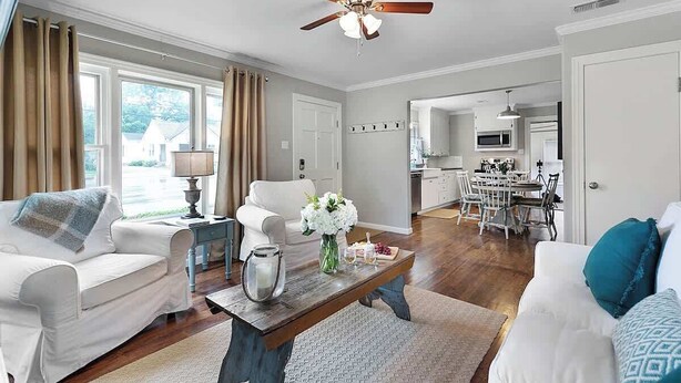 A spacious living room with a ceiling fan, a coffee table with flowers, a white sofa, and a dining area with a kitchen in the background. - New! Charming Home Just 4 Blks From Main St! (Fredericksburg)