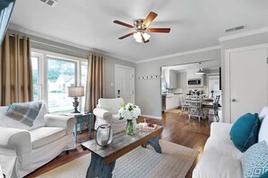 A spacious living room with a ceiling fan, a coffee table with flowers, a white sofa, and a dining area with a kitchen in the background. - New! Charming Home Just 4 Blks From Main St! (Fredericksburg)