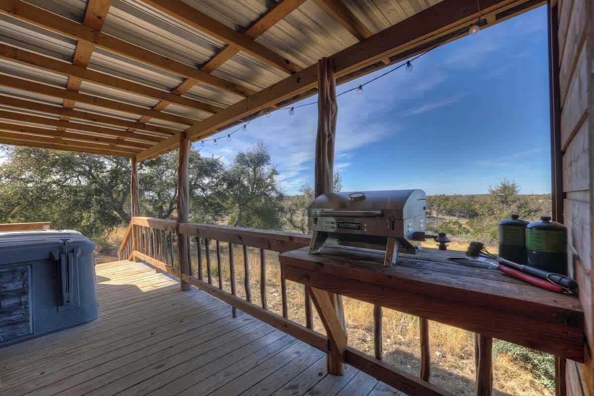 A covered outdoor deck with a grill, a hot tub, and a wooden railing.