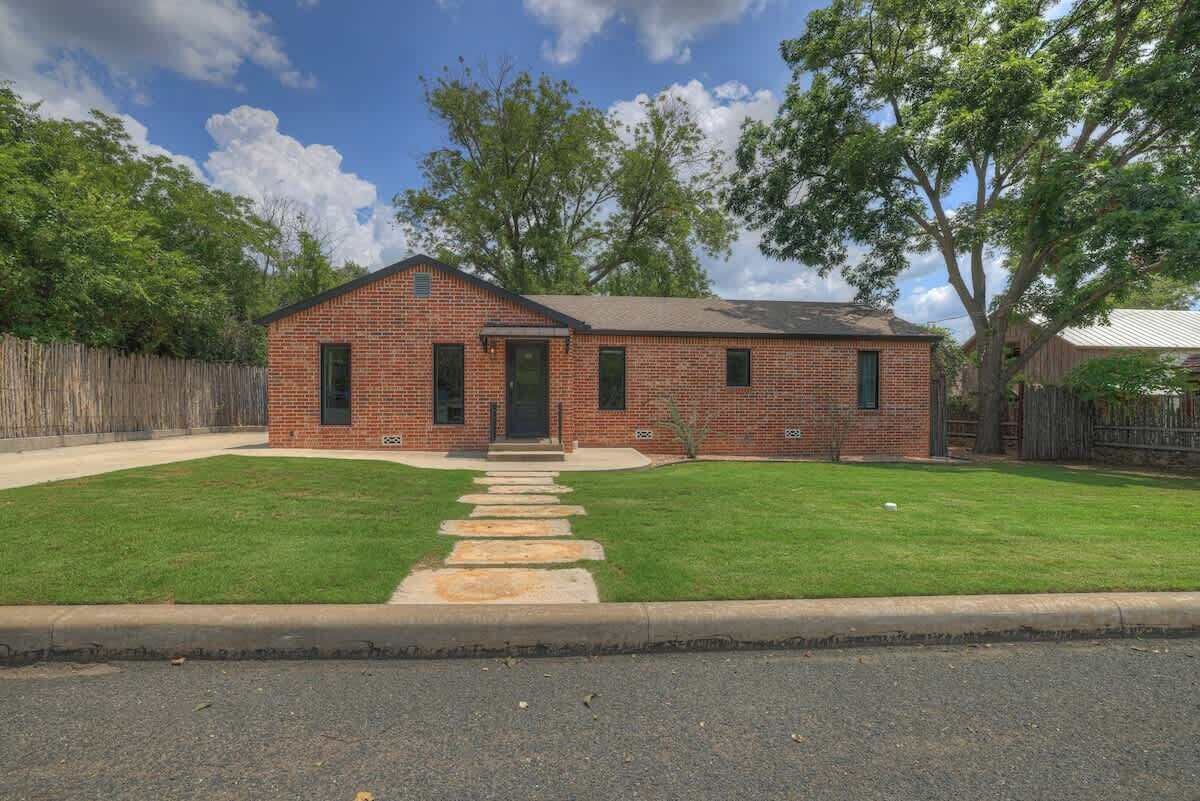 A single-story brick house with a front porch, a well-maintained lawn, and a wooden fence.