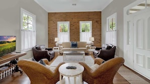 A living room with a sofa, two armchairs, a coffee table, and a TV. There are two windows with curtains, a wooden accent wall, and a white ceiling light.