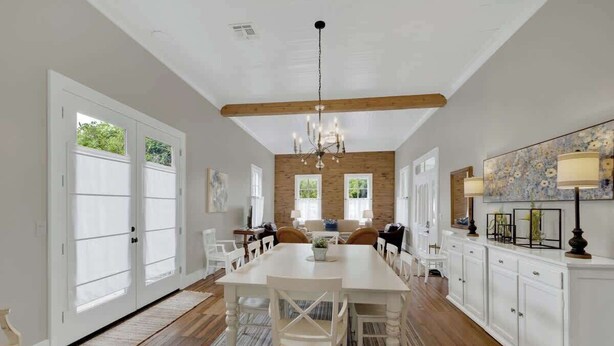 A spacious dining area with a long table, white chairs, and a chandelier. There are large windows on one side and a wooden ceiling beam.