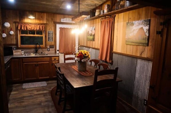 A rustic kitchen with wooden cabinets, a wooden dining table with chairs, and a vase of sunflowers. - " The Rusted Barn " (Edwards)