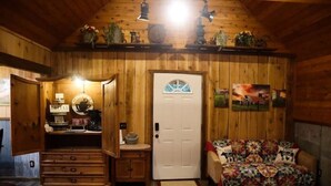 Una habitación acogedora con paredes de madera, una puerta blanca, un sofá floral, una mesita con una lámpara y un estante con adornos.
