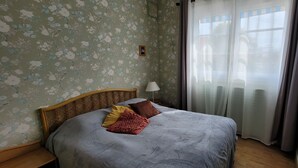 A bedroom with floral wallpaper, a wooden headboard, a bed with a grey bedspread and pillows, a bedside lamp, and a window with curtains. - A 5mn de la mer et du Bourg ,une Villa Plain Pied Vous Attend ! (Préfailles)