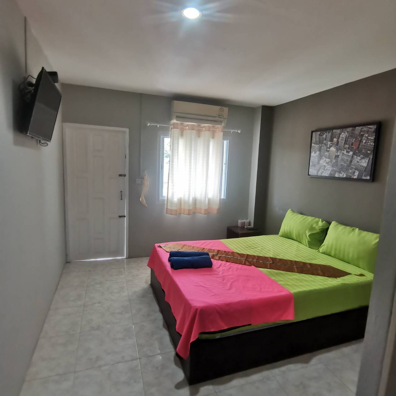 Een slaapkamer met een bed, een televisie, een raam met gordijnen en een stadsgezicht aan de muur.
