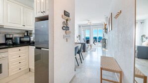 A modern kitchen with white cabinets, a stainless steel refrigerator, a microwave, and a coffee maker. There is a dining area with a table and chairs, and a view of the outdoors through large windows. - Sea Views 1 Bed Apt Colinas Del Faro (El Faro)