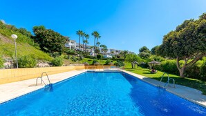 A swimming pool with clear blue water, surrounded by a landscaped garden with palm trees and a view of a hillside with buildings. - Sea Views 1 Bed Apt Colinas Del Faro (El Faro)