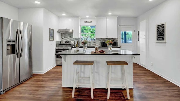 A modern kitchen with white cabinetry, a stainless steel refrigerator, and a central island with bar stools. - Gorgeous Bluebonnet House With Deck and Fire Pit (Fredericksburg)