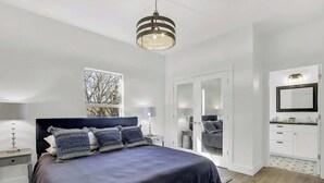 Un dormitorio con una cama grande, una mesita de noche, una lámpara colgante, una ventana con vistas y una puerta que da al baño.