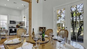 A dining area with a round table set for six, surrounded by wicker chairs. A wooden ceiling and a large window with a view of trees are visible.