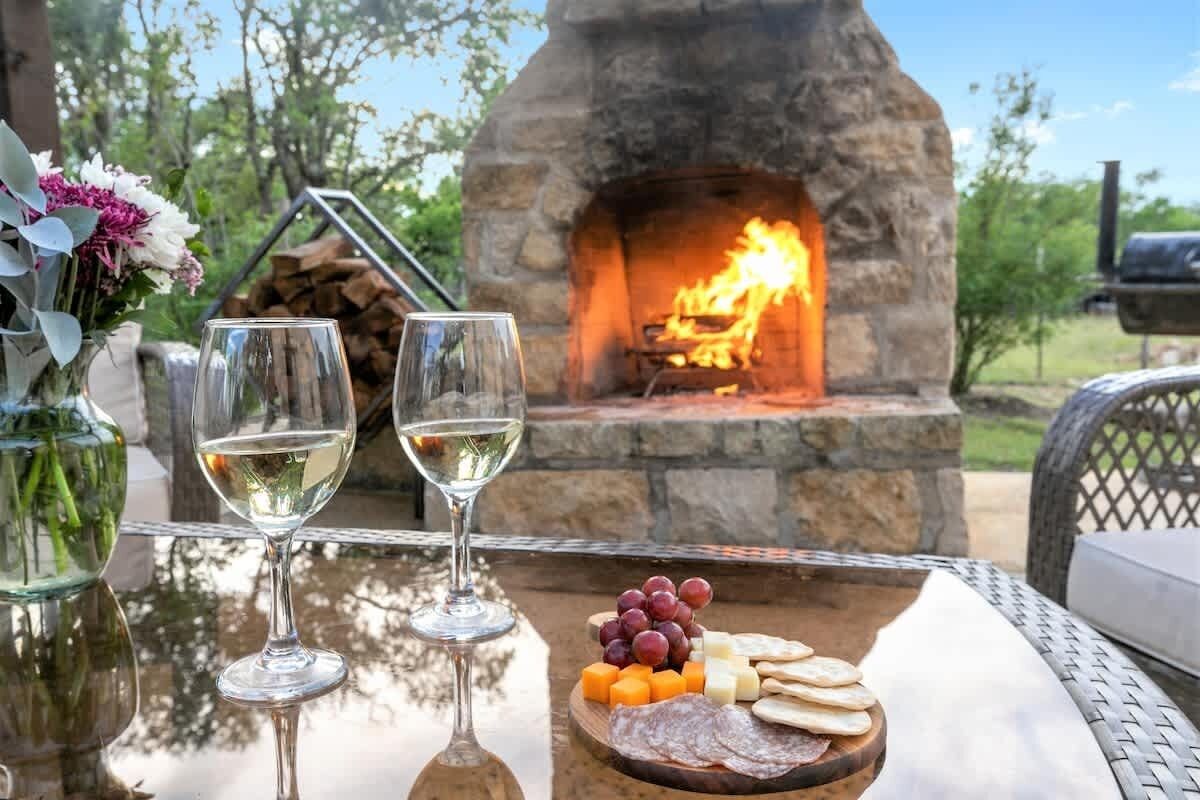 A stone fireplace with a fire, a glass table with two wine glasses and a cheese board with grapes and crackers.