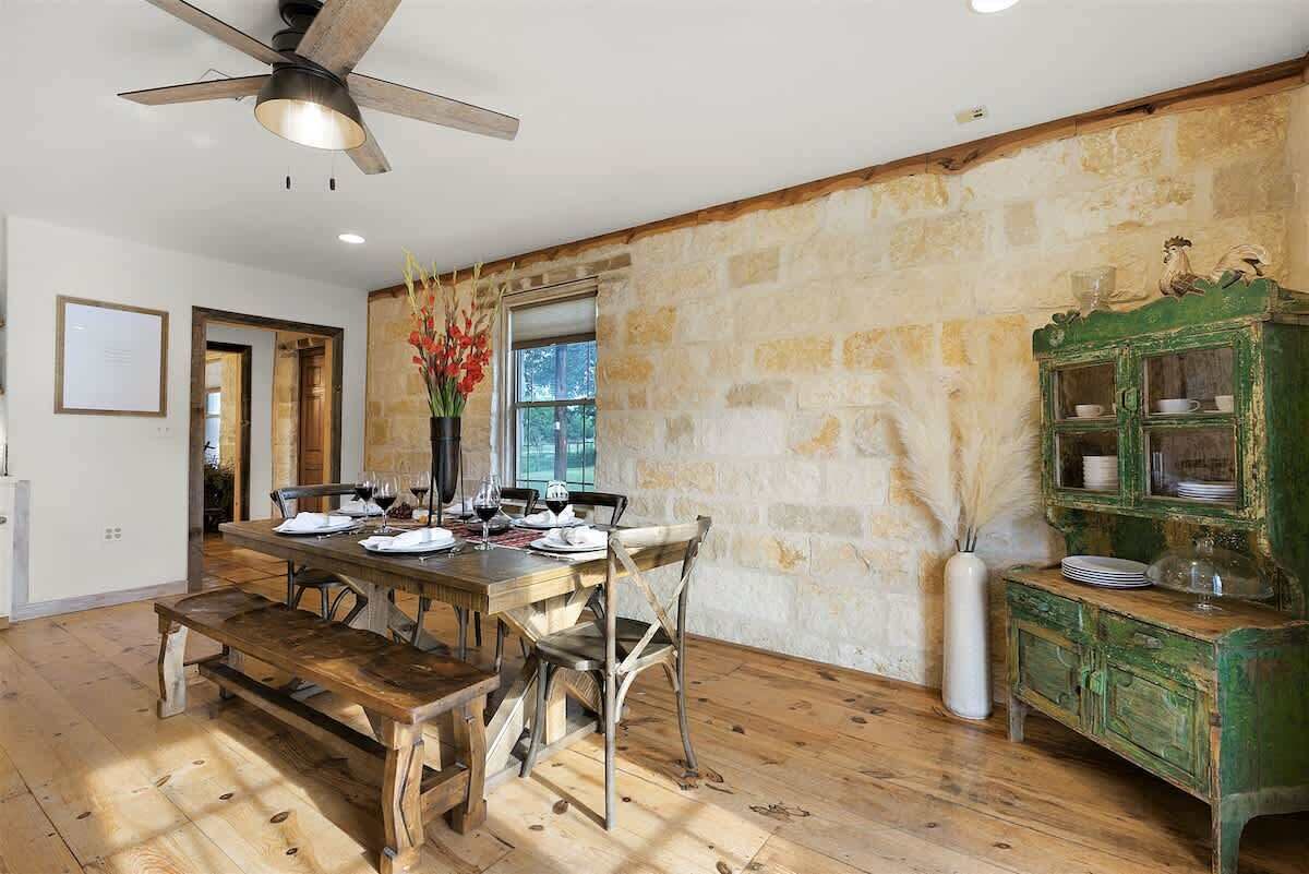 A dining area with a wooden table set for four, a ceiling fan, and a rustic stone wall.
