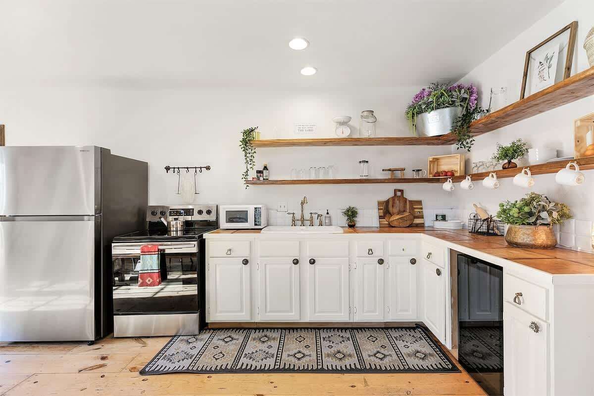 A modern kitchen with white cabinets, stainless steel appliances, wooden shelves, and a patterned rug.