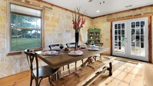 Une salle à manger avec une table en bois dressée pour quatre personnes, un vase rempli de fleurs rouges et une vue sur l’extérieur par une fenêtre.