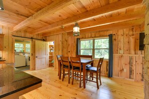 A wooden cabin interior with a dining table and chairs, a kitchen area, and a large window with curtains.