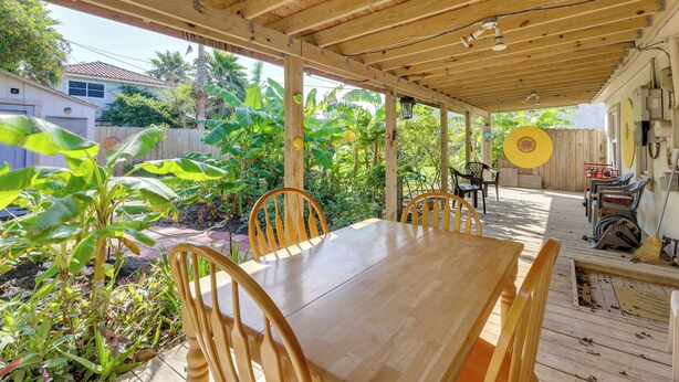 A covered patio with a wooden table and chairs, surrounded by lush greenery and a wooden fence.