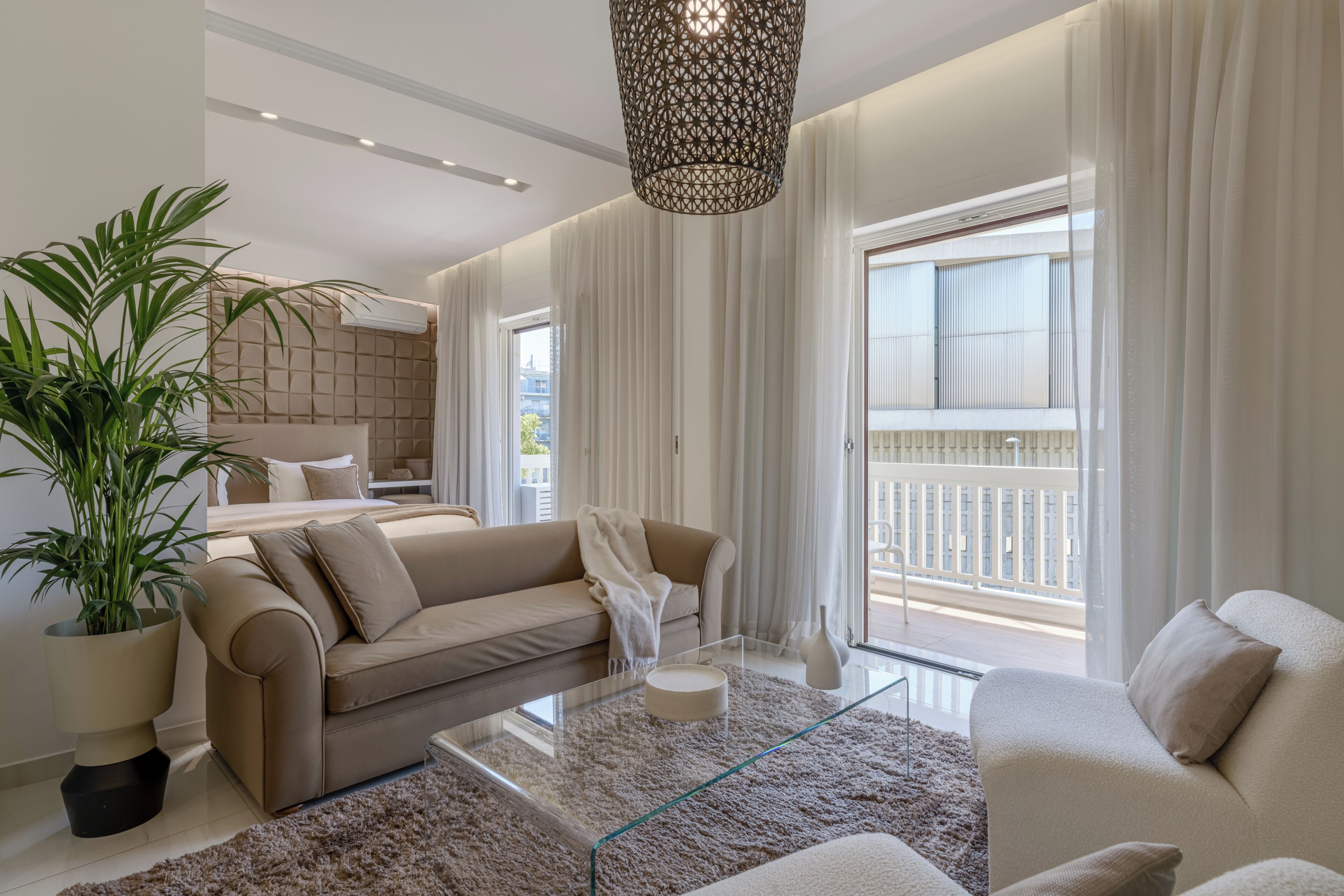 En moderne stue med et glasbord, beige sofaer, en stor plantepotte og en balkon med gelænder.
