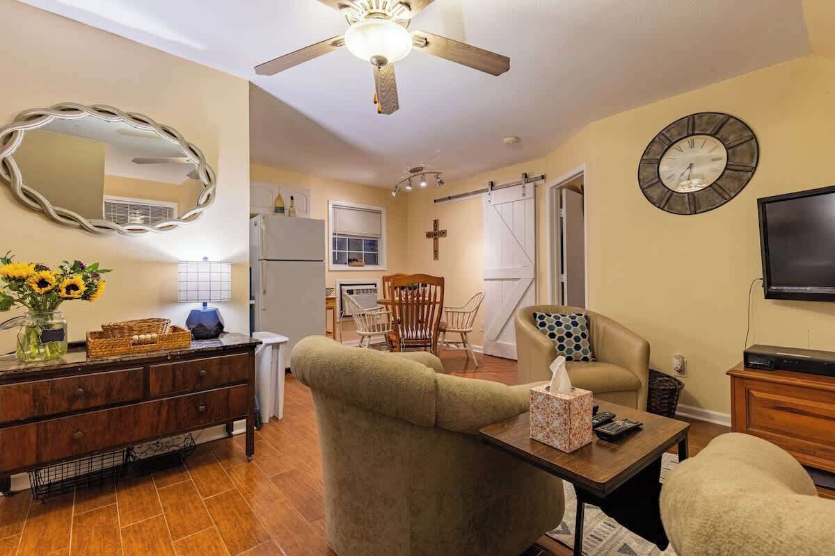 Un salon avec un canapé, une table basse, un téléviseur, un ventilateur de plafond, une horloge et, au fond, un coin cuisine.