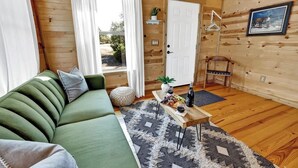 A cozy wooden cabin interior with a green sofa, a wooden coffee table with fruits and a bottle, and a view of the outdoors through a window. - Rocky Ridge Cedar Cabin With Hot Tub & Amazing Views (Fredericksburg)