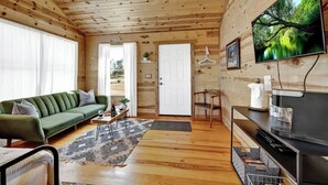 A wooden cabin interior with a sofa, television, and a door leading outside. - Rocky Ridge Cedar Cabin With Hot Tub & Amazing Views (Fredericksburg)