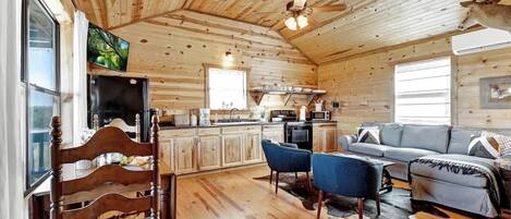 A rustic cabin interior with a wooden ceiling, a dining area with a wooden table and chairs, a kitchen with modern appliances, and a living area with a sofa and a mounted deer head.