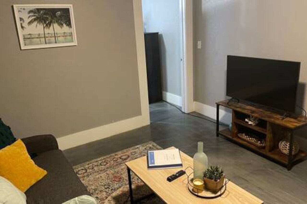 A living room with a sofa, a coffee table with a remote control, a TV on a wooden stand, and a door leading to another room.