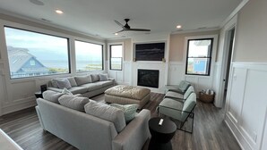 A spacious living room with a sectional sofa, a fireplace, and large windows offering a view of the ocean. - 6BR - 6.5 Bath - Sleeps 14-Amazing Sunsets, Creating Lifelong Memories Together  (Salvo)