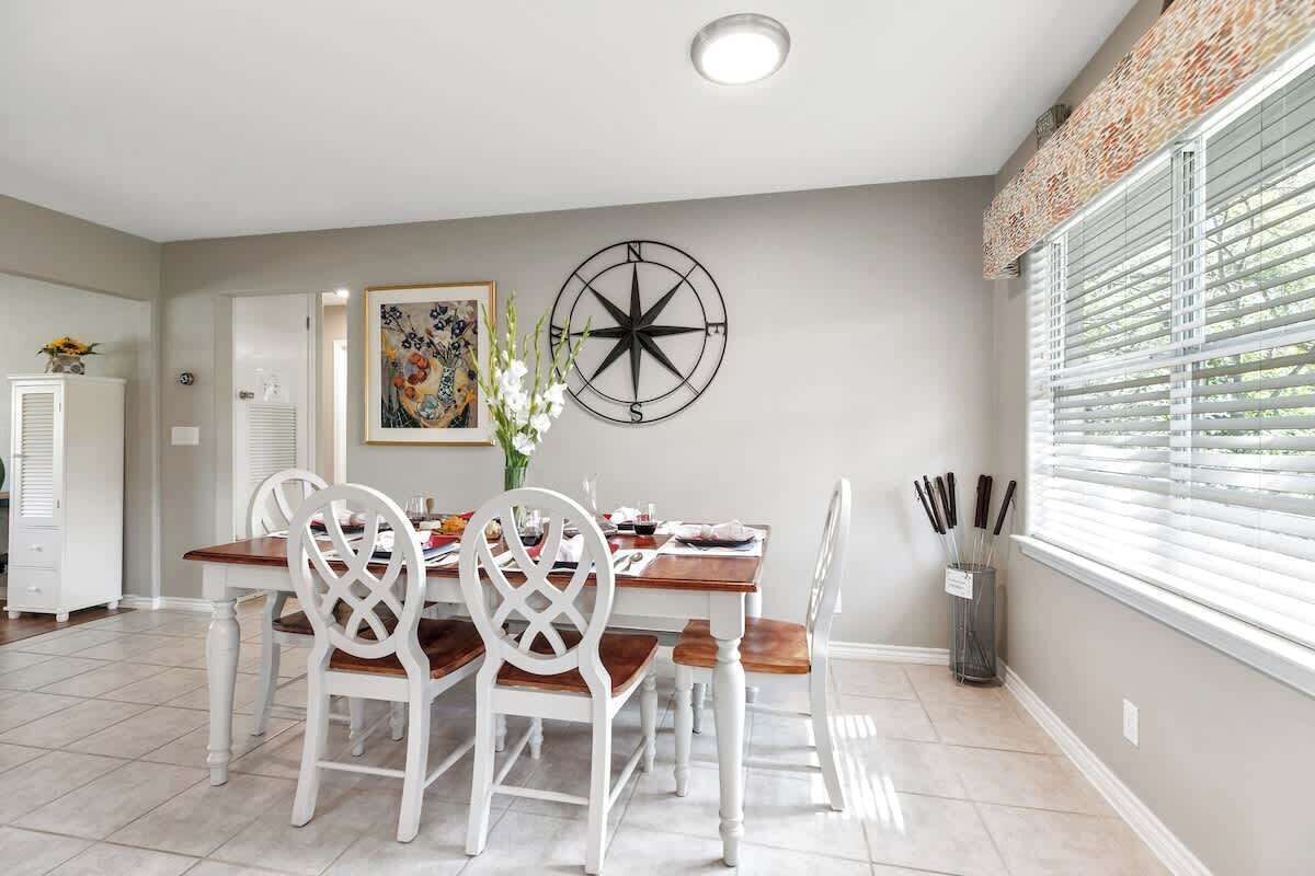 A dining area with a wooden table and white chairs, a compass wall decoration, and a window with blinds.