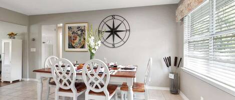 A dining area with a wooden table and white chairs, a compass wall decoration, and a window with blinds.