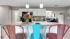 A modern kitchen with granite countertops, white cabinets, and a central island with two chairs. There are hanging pendant lights, a refrigerator, and a coffee machine.