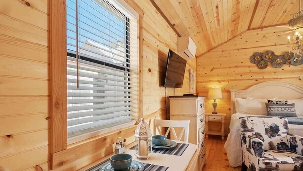 Un acogedor interior de cabaña con una mesa de comedor lista para dos personas, una cama con ropa de cama floral y un televisor montado.