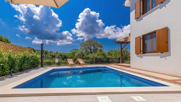 Ein rechteckiger Pool mit gefliestem Rand, umgeben von einer Betonplattform. Im Hintergrund sind zwei Liegestühle und eine Pergola zu sehen.