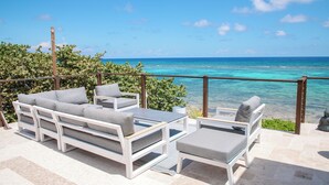 A terrace with a white sofa and chairs, overlooking a turquoise sea.