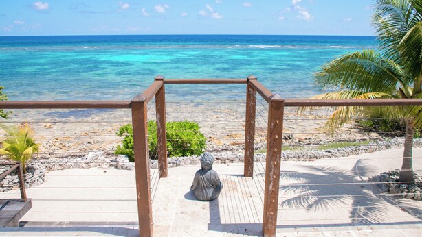 A wooden deck with a Buddha statue, overlooking a beach with clear turquoise waters and a palm tree.