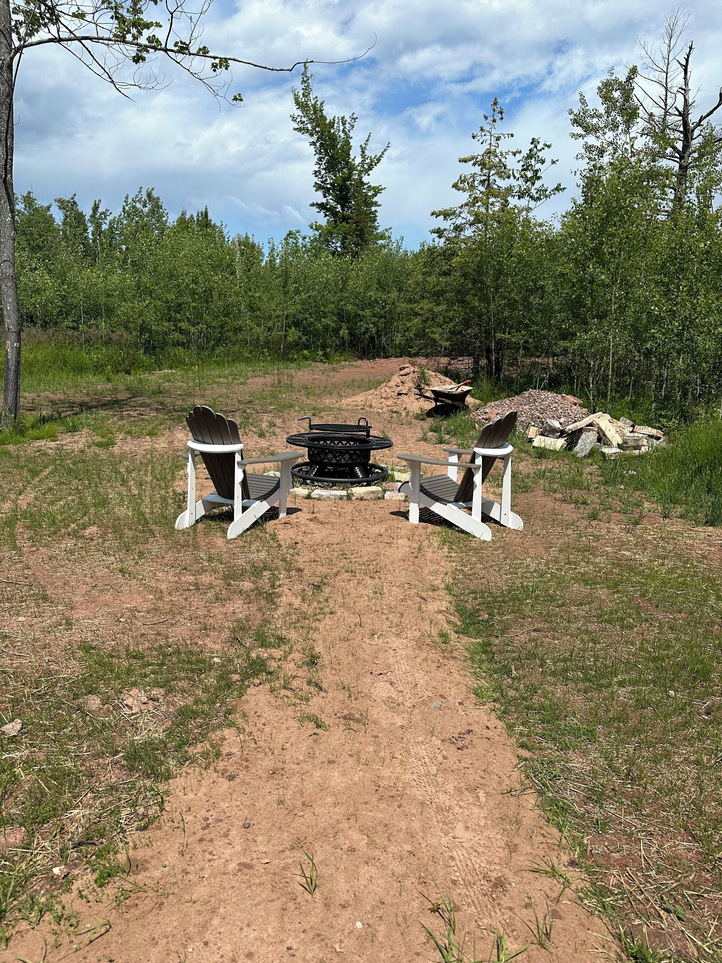 A campsite with two Adirondack chairs facing a central fire pit, surrounded by a dirt path and wooded area.